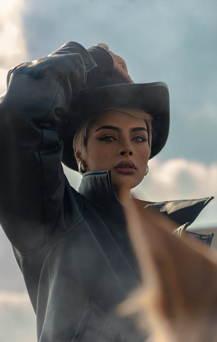 A confident woman wearing a leather jacket and cowboy hat poses dramatically against an outdoor sky backdrop.